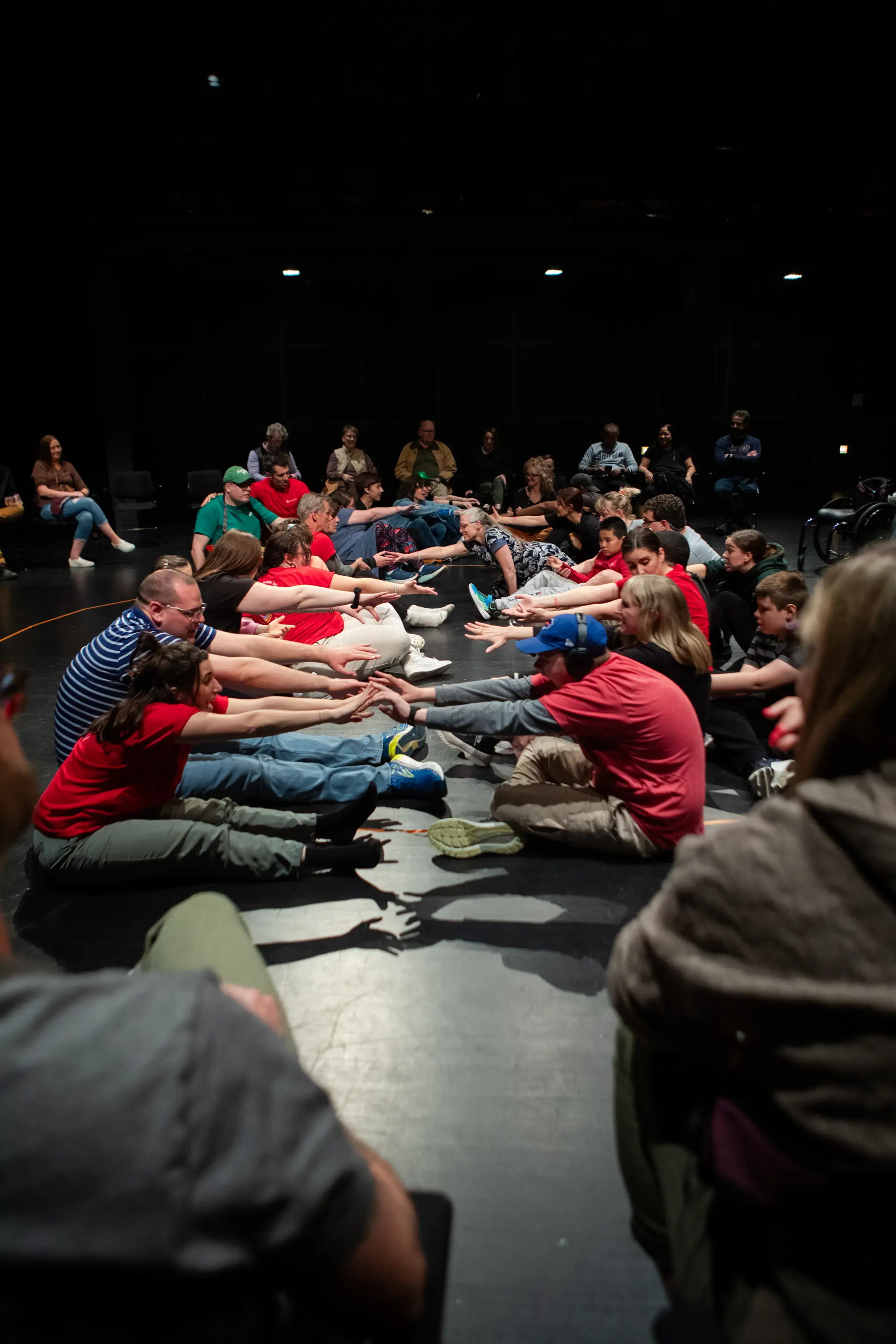 A group of autistic participants, families, and facilitators sitting in a large circle on stage, reaching their arms forward together during an interactive Flute Theatre workshop at the Wexner Center for the Arts.