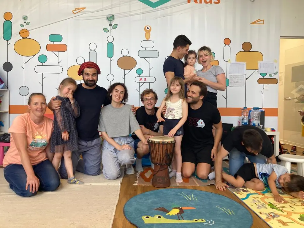A warm group photo of performers and children in a colorful playroom, with a drum and playful wall designs, creating a joyful and welcoming atmosphere.