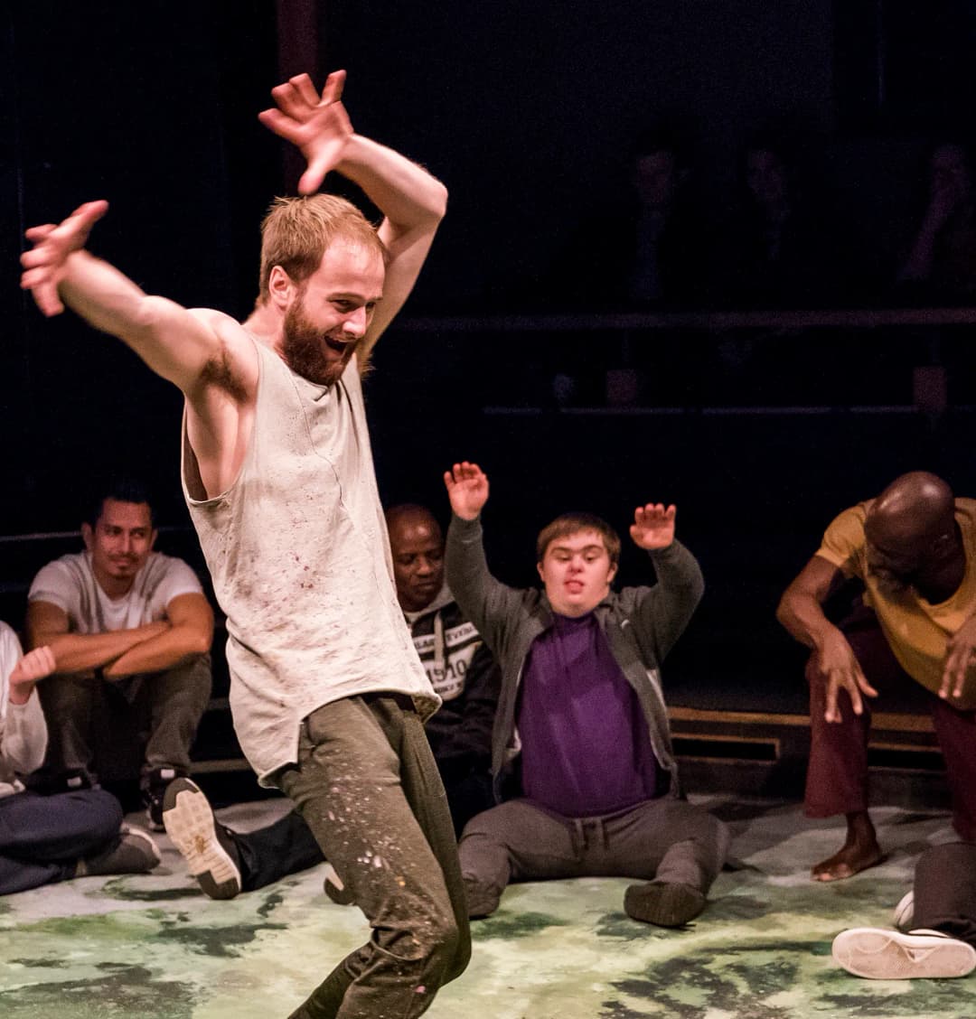 A man in a white tank top energetically dances with arms raised, while participants sitting around him mimic his movements with smiles and engagement.